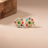 A pair of gold-toned stud earrings with colorful gemstone accents and white pearl-like details, displayed on a warm beige surface with a blurred wooden block in the background.