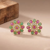 A pair of gold-plated floral stud earrings with pink and green gemstones, displayed on a beige surface with a wooden block in the background.
