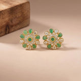 A pair of gold floral stud earrings with green stones and clear stones, displayed on a beige surface with a wooden block in the background.