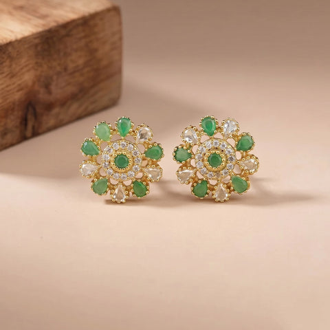 A pair of gold floral stud earrings with green stones and clear stones, displayed on a beige surface with a wooden block in the background.