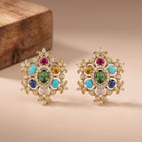 A pair of ornate gold-toned statement earrings with multicolored gemstones, including green, turquoise, pink, blue, and amber, accented with clear crystals, displayed on a beige surface with a wooden block in the background.