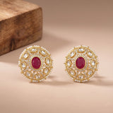 A pair of ornate gold-toned stud earrings with oval red center stones and surrounding clear stones set in a pearl-like border, displayed on a warm beige surface with a wooden block in the background.