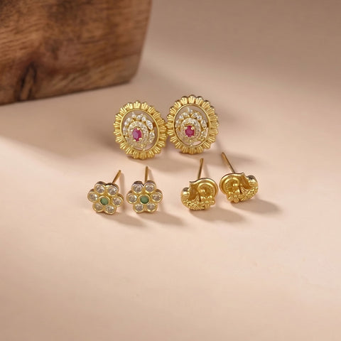 A pair of round gold-toned stud earrings with a central pink stone and a set of four smaller flower-shaped gold-toned stud earrings with green and clear stones, displayed on a beige surface.