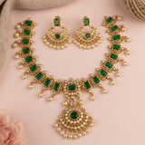 Gold-plated Indian jewelry set featuring a necklace, earrings, and a maang tikka, decorated with emerald-cut green stones and white faux pearls, displayed on a soft pink background.