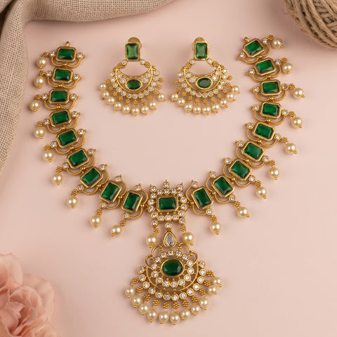 Gold-plated Indian jewelry set featuring a necklace, earrings, and a maang tikka, decorated with emerald-cut green stones and white faux pearls, displayed on a soft pink background.