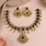 Gold-plated Indian ethnic jewelry set featuring a necklace and matching earrings with dark blue rectangular stones and white faux pearls, arranged on a soft pink background with a beige fabric corner.