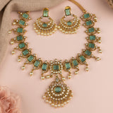 Gold-plated Indian ethnic jewelry set featuring a necklace, earrings, and a matching bracelet, embellished with rectangular green stones and small white faux pearls, displayed on a soft pink background.