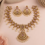 Gold-toned traditional Indian jewelry set featuring a necklace, earrings, and bangles with lavender-pink rectangular stones and white faux pearls, arranged on a soft pink background with decorative fabric and a flower.