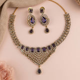 Gold-toned traditional Indian jewelry set featuring a statement necklace and matching drop earrings, embellished with clear crystals and purple gemstones, displayed on a soft pink background.