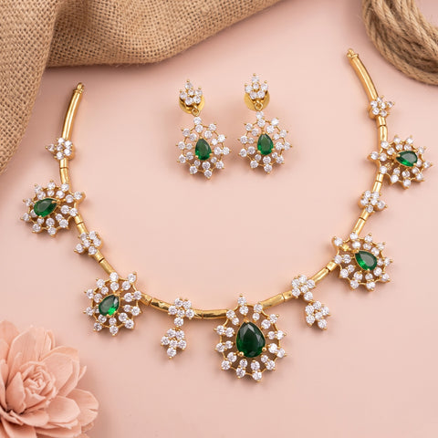 Gold-toned jewelry set with a necklace and matching earrings, featuring green teardrop gemstones surrounded by clear crystal-like stones, arranged on a soft pink background with burlap and a rose prop.
