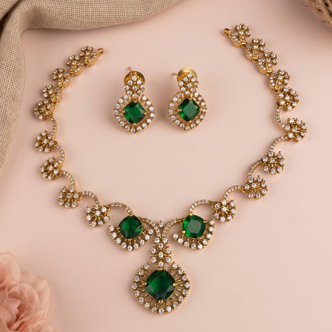 Gold-tone jewelry set with a necklace and matching earrings, featuring green gemstone centers surrounded by clear rhinestones, arranged on a soft pink background with a beige fabric accent.