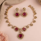 Gold-plated jewelry set featuring a necklace and matching earrings with large square pinkish-red gemstones surrounded by clear rhinestones, displayed on a soft pink background with decorative fabric.