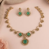Gold-toned jewelry set featuring a necklace and matching earrings with oval green stone centers surrounded by small clear stones, arranged on a soft pink background.