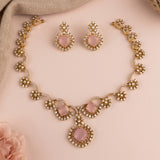 Gold-toned jewelry set with a necklace, matching earrings, and a bracelet, featuring large pink stones surrounded by clear rhinestones, arranged on a soft pink background.