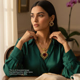Woman wearing a green blouse and gold necklace with a red gemstone, sitting indoors.
