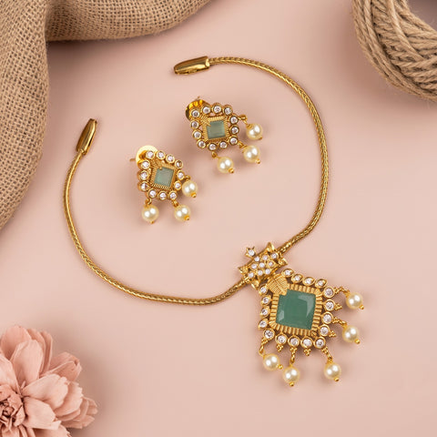 Gold-toned jewelry set with a necklace, earrings, and a decorative pendant, arranged on a soft pink background with burlap and a pink flower prop.