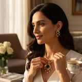 Woman wearing a gold necklace with a green gemstone, holding a cup of tea in a cozy room.