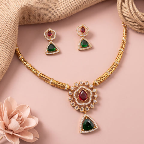 Gold-toned traditional jewelry set featuring a necklace and matching earrings with green teardrop gemstones and red oval stones, accented with small clear stones, arranged on a pink background with burlap and a pale flower prop.