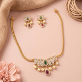 Gold-toned traditional jewelry set featuring a necklace and matching earrings, decorated with white stones, green teardrop gems, and red teardrop gems, styled on a pink background with burlap and a pink flower.