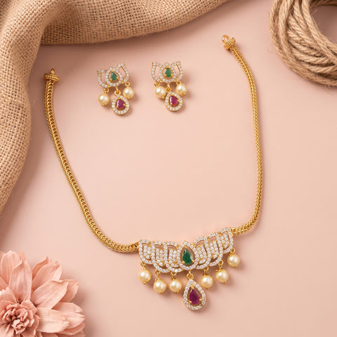 Gold-toned traditional jewelry set featuring a necklace and matching earrings, decorated with white stones, green teardrop gems, and red teardrop gems, styled on a pink background with burlap and a pink flower.
