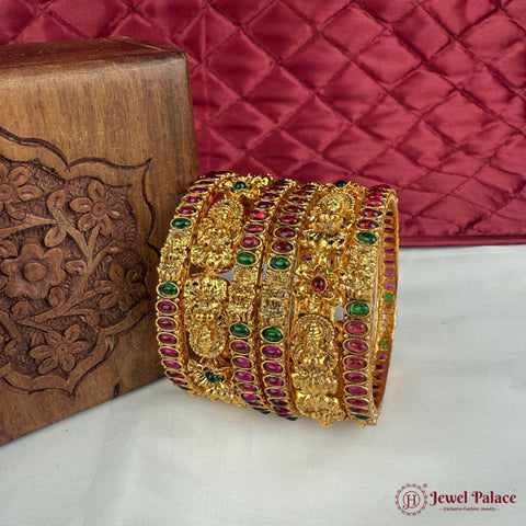 Gold bangles with gemstones on a white surface next to a wooden box, with a red quilted fabric background.