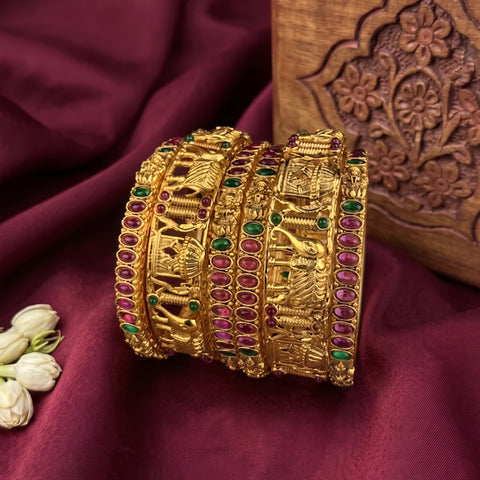 A set of five traditional Indian gold bangles (kara) decorated with detailed engravings and accented with pink and green stones, displayed on a draped maroon fabric background.