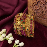 A stack of ornate gold bangles decorated with red and green stones, displayed on a deep burgundy velvet fabric with white flowers and a carved wooden block in the background.