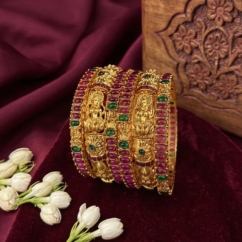 A stack of ornate gold bangles decorated with red and green stones, displayed on a deep burgundy velvet fabric with white flowers and a carved wooden block in the background.
