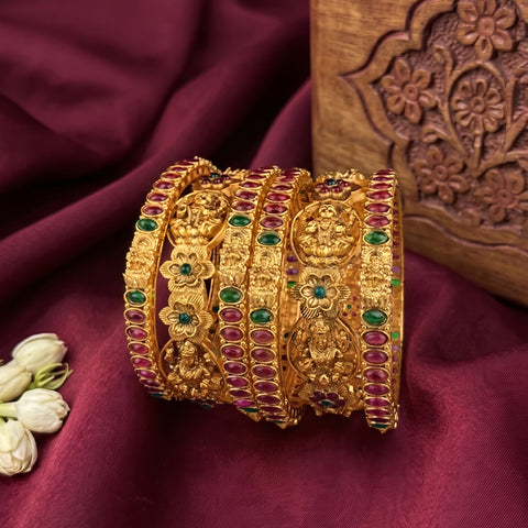 A set of five ornate gold bangles decorated with red and green stones and detailed motifs, arranged on a draped burgundy fabric next to white jasmine flowers and a carved wooden block.