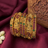 A set of ornate gold bangles decorated with red and green stones, arranged on a deep burgundy fabric backdrop next to a carved wooden block.