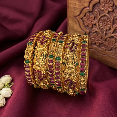 A set of ornate gold bangles decorated with red and green stones, arranged on a deep burgundy fabric backdrop next to a carved wooden block.