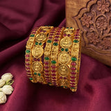 A set of five wide gold bangles decorated with round and oval red and green stones, displayed on a deep burgundy satin fabric next to a carved wooden block.