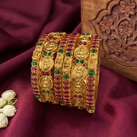 A set of five wide gold bangles decorated with round and oval red and green stones, displayed on a deep burgundy satin fabric next to a carved wooden block.