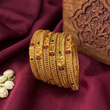 A stack of gold bangles with red and green stone accents, arranged on a deep burgundy fabric backdrop next to a carved wooden box and white flowers.