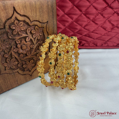 Gold bracelet with green and red stones on a white surface with a wooden box and red fabric in the background.