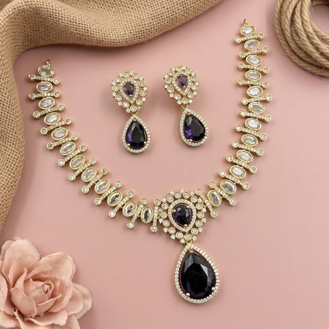 Gold-toned traditional jewelry set featuring a necklace, earrings, and matching bangles with clear stones and dark purple teardrop gemstones, arranged on a pink background with burlap and a rose prop.