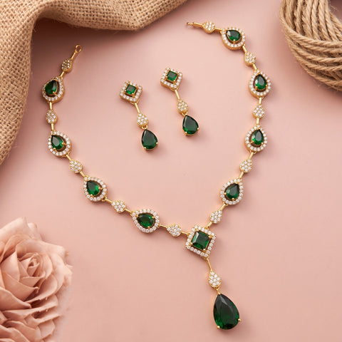Gold jewelry set with emerald green stones and white crystal accents, including a necklace, matching earrings, and a bracelet, arranged on a pink background with decorative burlap and a rose.