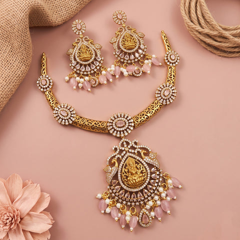 Gold-toned traditional jewelry set with a necklace, earrings, and pendant featuring pink stone drops and white faux pearls, arranged on a soft pink background with burlap and a flower prop.