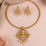 Gold-toned traditional jewelry set featuring a braided necklace with an ornate deity pendant, matching drop earrings, and pearl accents, arranged on a soft pink background with a beige cloth and pale pink flower.