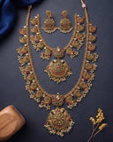 An ornate gold-toned traditional necklace set with matching earrings, decorated with red and green enamel work and white pearl-like beads, arranged on a dark navy background with decorative props.