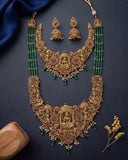 An ornate gold-toned traditional necklace set with matching earrings, decorated with green bead strands, red and green stones, and small white stones, arranged on a dark navy background with decorative fabric and dried flowers.