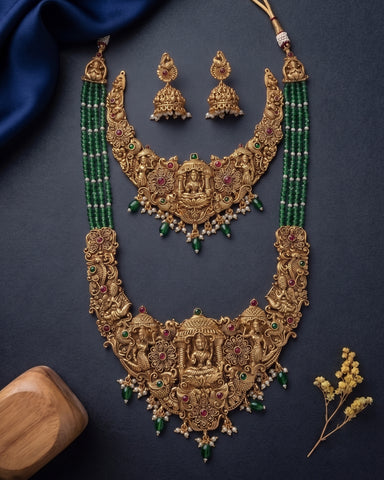 An ornate gold-toned traditional necklace set with matching earrings, decorated with green bead strands, red and green stones, and small white stones, arranged on a dark navy background with decorative fabric and dried flowers.