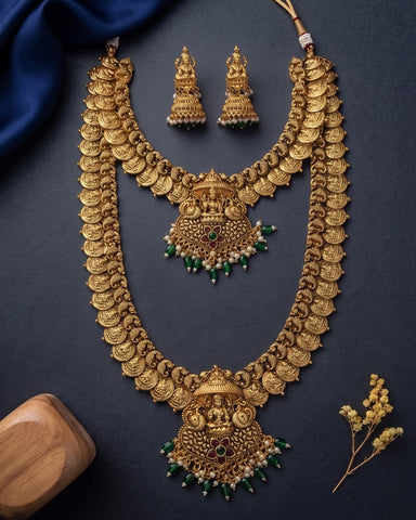 An ornate gold-toned traditional necklace set with matching earrings, arranged on a dark navy background with decorative blue fabric, a wooden block, and small dried flowers.