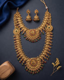 An ornate gold-toned traditional necklace set with matching earrings, arranged on a dark navy background with a blue fabric drape, a wooden block, and small dried flowers.