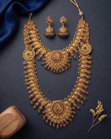 An ornate gold-toned traditional necklace set with matching earrings, arranged on a dark navy background with a blue fabric drape, a wooden block, and small dried flowers.