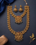 An ornate gold-toned traditional necklace set with matching earrings, featuring intricate filigree detailing, red gemstone accents, and dangling dark green bead clusters, arranged on a dark navy background with decorative fabric and dried flowers.