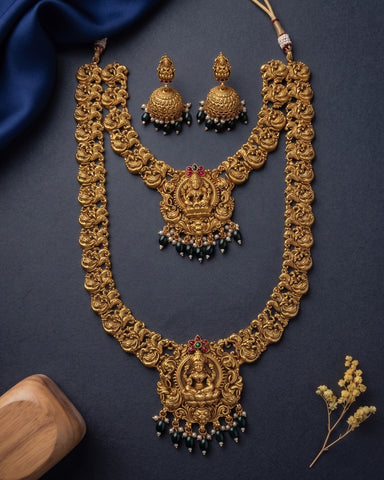 An ornate gold-toned traditional necklace set with matching earrings, featuring intricate filigree detailing, red gemstone accents, and dangling dark green bead clusters, arranged on a dark navy background with decorative fabric and dried flowers.