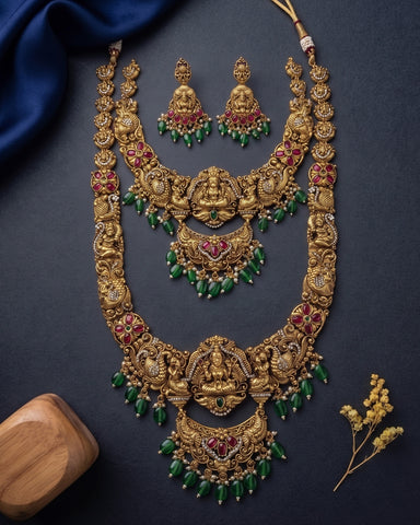 An ornate gold-toned traditional Indian jewelry set featuring a necklace, matching earrings, and a pendant with red stone accents and green bead drops, arranged on a dark navy background with decorative fabric and a small wooden block.