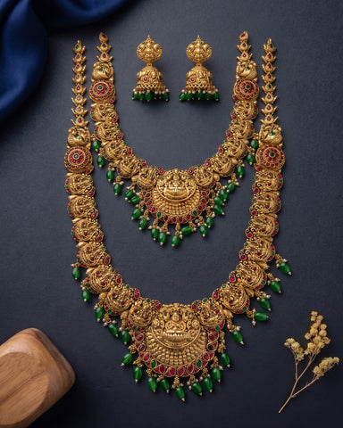 An ornate gold-toned traditional jewelry set laid out on a dark navy background, including a large necklace, matching earrings, and a smaller necklace, decorated with green bead drops and red stone accents. A small wooden block and dried floral stems are placed as styling props.
