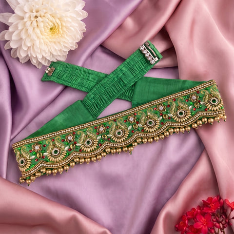 A green fabric belt with ornate gold embroidery, bead borders, and small red and green stone accents, laid flat on draped pink satin fabric with decorative flowers.